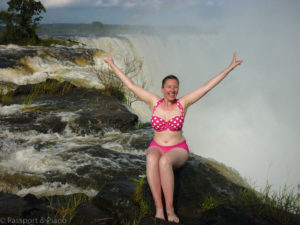 An image of me with my arms in the air sat on the edge of Victoria Falls with the water and mist behind.